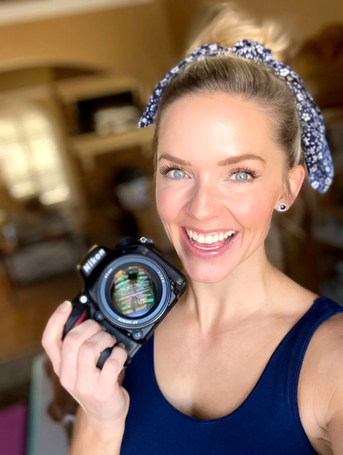 A woman with blonde hair, blue eyes, and a blue and white patterned headscarf, smiling and holding a Nikon camera in a cozy, wood-paneled room.