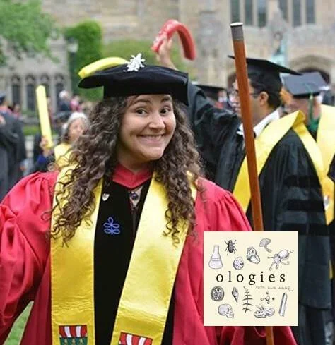 A woman in a red graduation gown and yellow stole, smiling at a graduation ceremony with other graduates in black caps and gowns in the background.