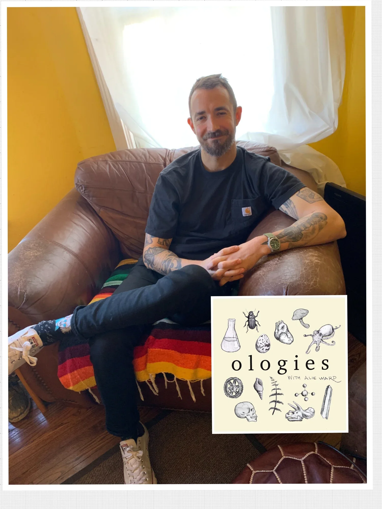 A man with tattoos sitting on a brown leather couch, wearing a black t-shirt, black pants, and white sneakers. The background features a yellow wall and a white curtain covering a window. There is a colorful striped blanket on the couch and a book or magazine titled 'Ologies with Alie Ward' with illustrated scientific and biological symbols in the bottom right corner.