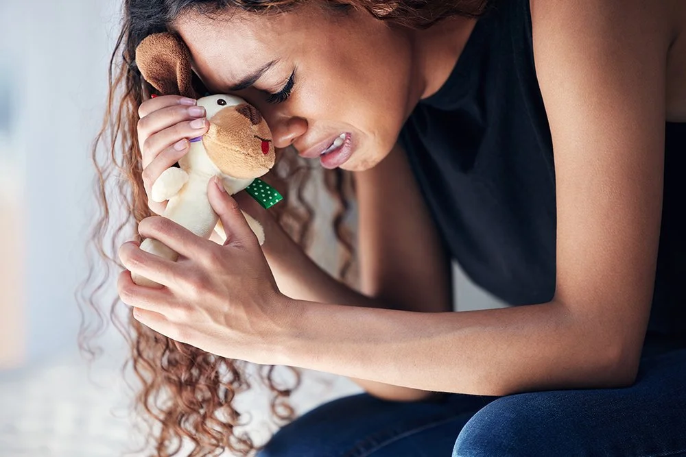 A woman with curly hair and a black shirt crying, holding a stuffed dog close to her face.