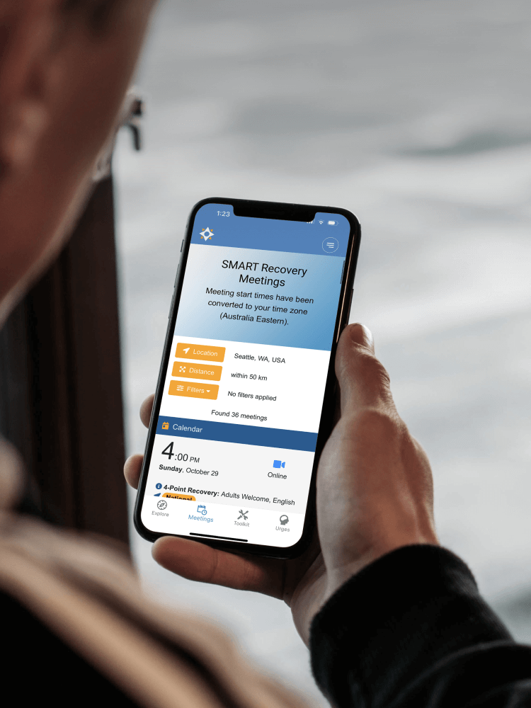 Person holding a smartphone displaying the SMART Recovery Meetings app, with information about local meetings, filters, and schedule.