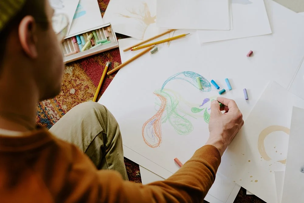 An artist drawing with pastel chalks on a large sheet of paper, creating a colorful abstract design. Art supplies and sketches are scattered on the floor around them.