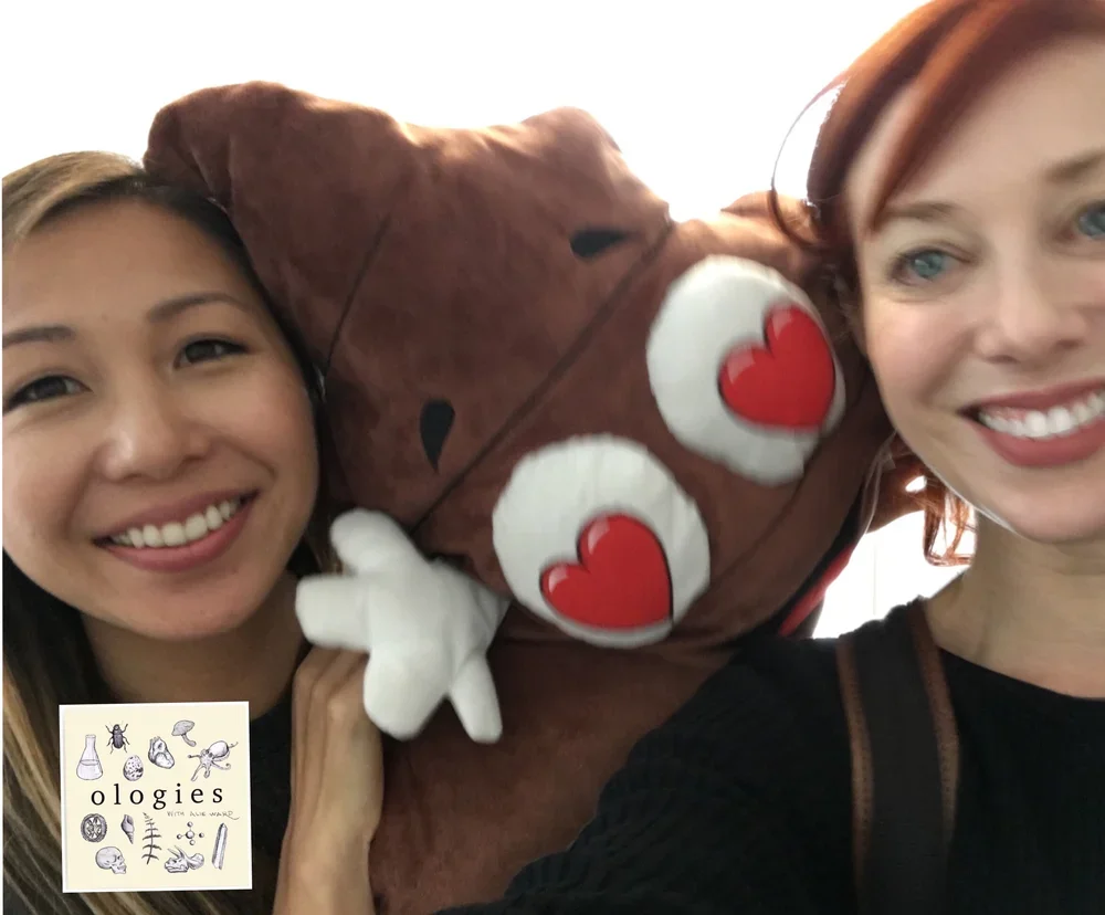 Two women smiling, holding a large plush poop emoji with heart eyes between them.