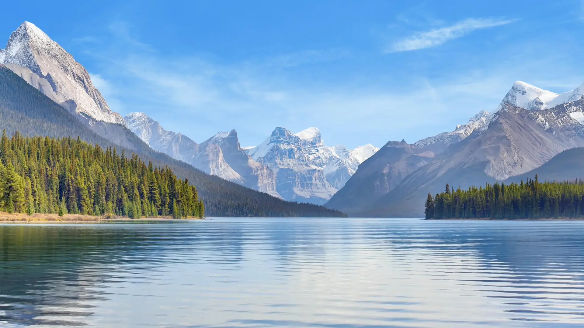 Scenic view of a mountain landscape with snow-capped peaks, a calm lake in the foreground, and evergreen trees along the shoreline under a blue sky with some clouds.