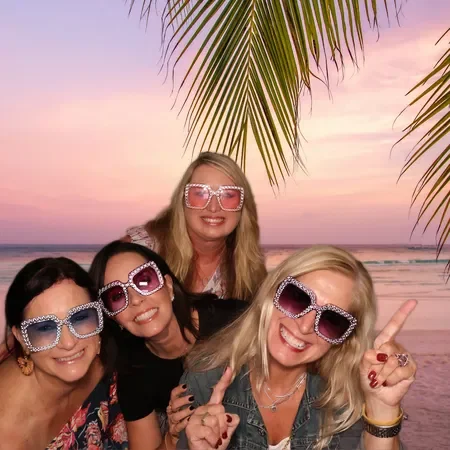 Four women smiling and wearing sunglasses on a beach at sunset, with palm tree leaves overhead.
