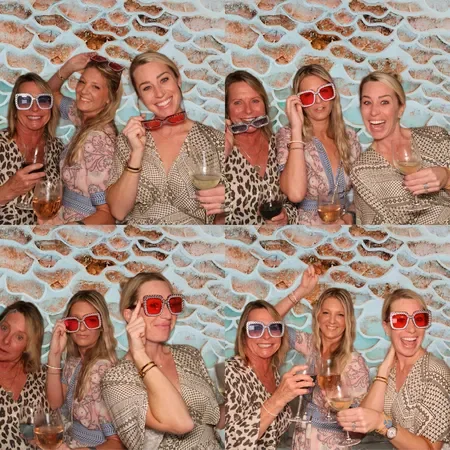 Six women at a party wearing sunglasses, holding drinks, and smiling in front of a decorative brick wall.