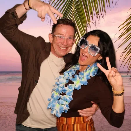 Two people smiling and making peace signs at the beach during sunset, with palm trees in the background, dressed in casual beach attire with accessories.