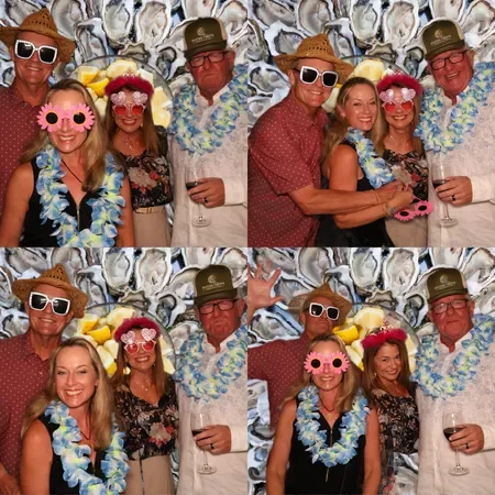 Four people posing for a photo booth picture with a floral backdrop, wearing tropical accessories like leis, straw hats, sunglasses, and pink flower glasses, at a tropical-themed party.