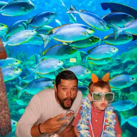 A man and a girl wearing sunglasses and a colorful lei, posing with a background of blue and yellow fish in an aquarium