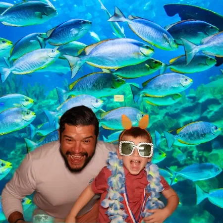 A man and a child posing in front of an aquarium filled with colorful fish. The child is wearing sunglasses, a flower lei, and a headband with bunny ears.