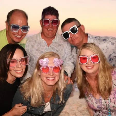 Group of six friends wearing colorful, sparkly sunglasses, smiling together at sunset