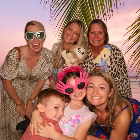 Group of five women and a boy smiling with tropical sunset background and palm leaves, some wearing playful glasses and holding plush toys.