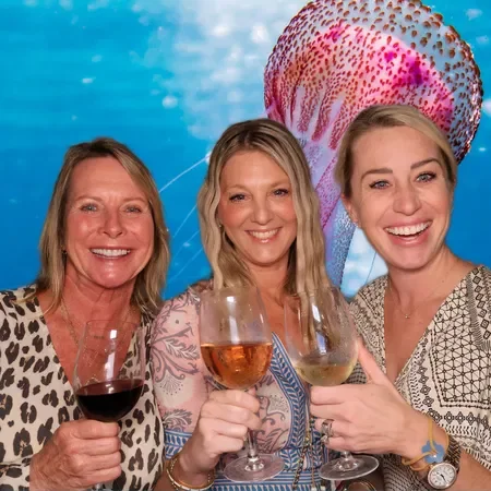 Three women smiling and holding wine glasses at a social gathering, with a colorful octopus underwater backdrop.