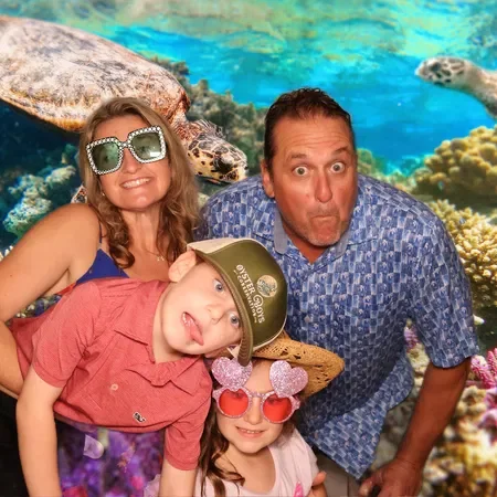 A family of four posing in front of an ocean-themed backdrop with a turtle and coral. The mother wears large sunglasses, the father makes a surprised face, the boy wears a green hat and sticks his tongue out, and the girl wears pink sunglasses with h