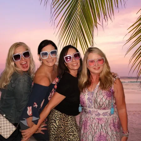 Four women in casual summer outfits and colorful sunglasses smiling at the beach during sunset with palm trees overhead.