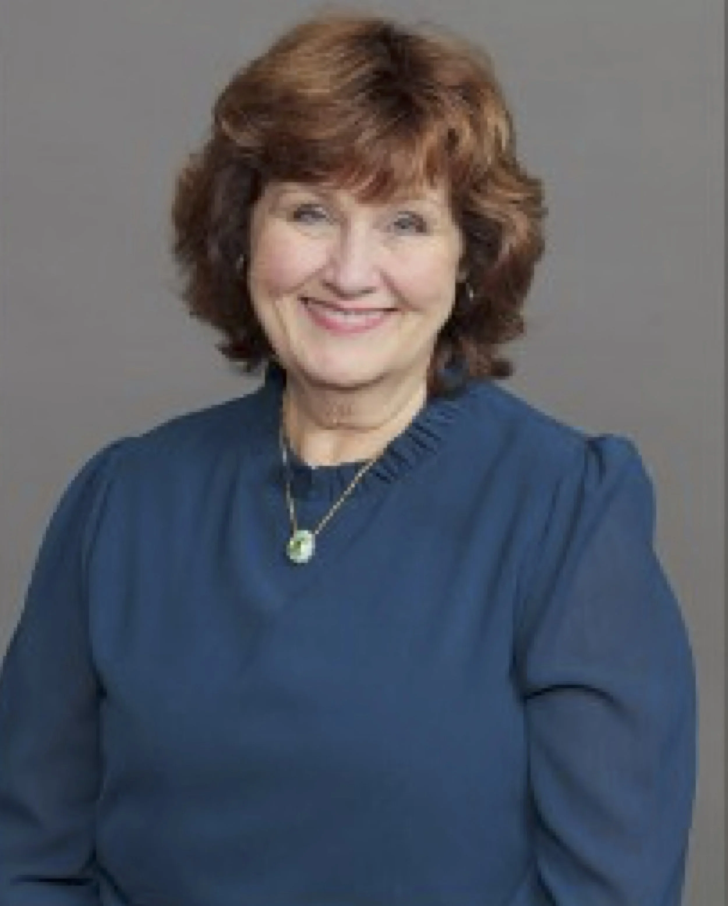 A woman with short, wavy brown hair, smiling, wearing a blue top and a silver necklace with a green pendant, against a plain gray background.