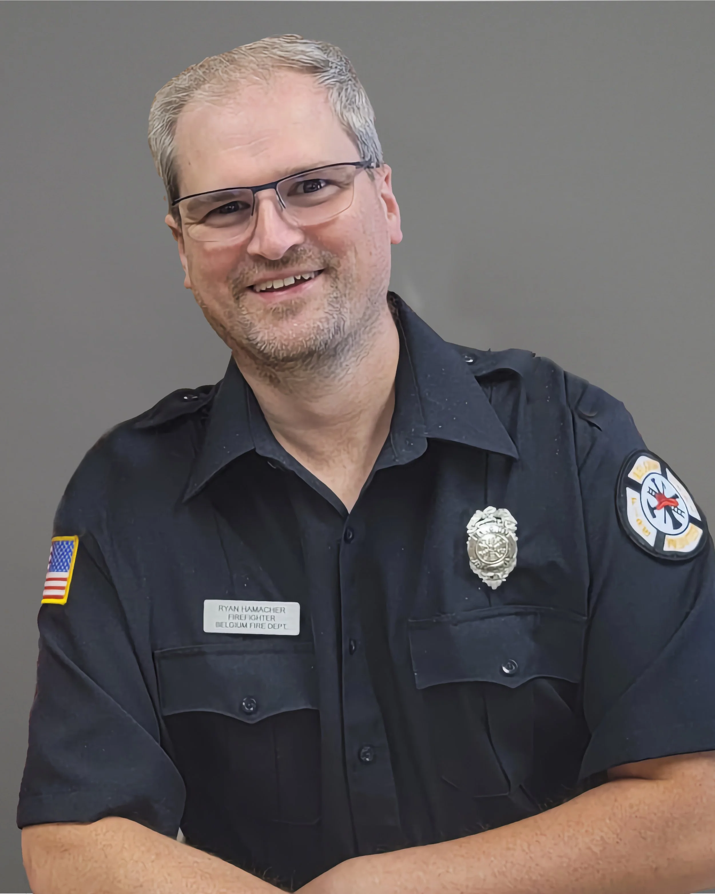 A smiling male firefighter wearing glasses and a black uniform with patches and a badge, sitting against a plain background.