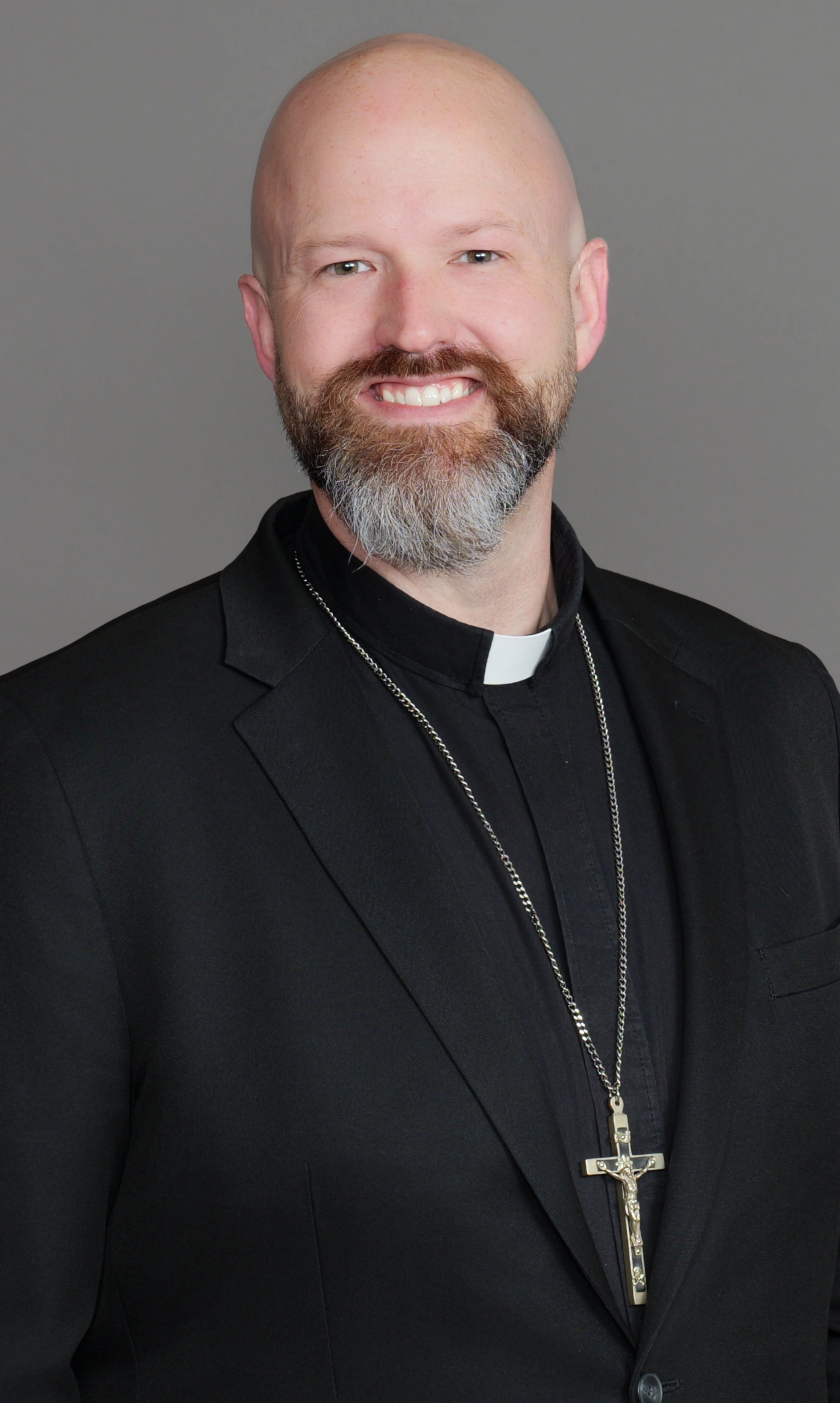 A picture of Pastor Lewis Polzin wearing a black suit, a clerical collar, and a silver crucifix necklace, posing against a plain gray background.