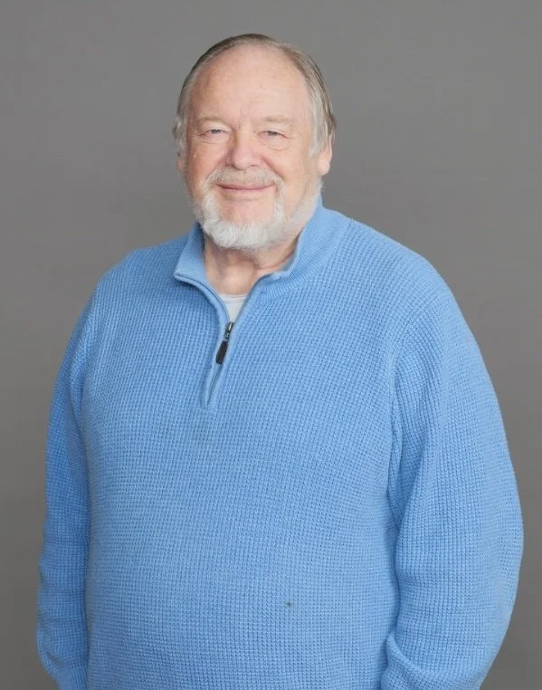Portrait of a smiling older man with gray hair and beard, wearing a blue zip-up sweater, against a gray background.