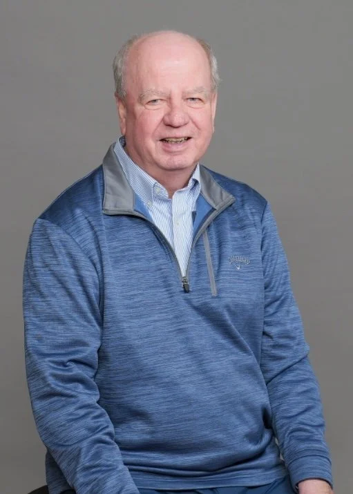 Portrait of an older man with a friendly expression, wearing a blue jacket and a collared shirt, against a plain gray background.