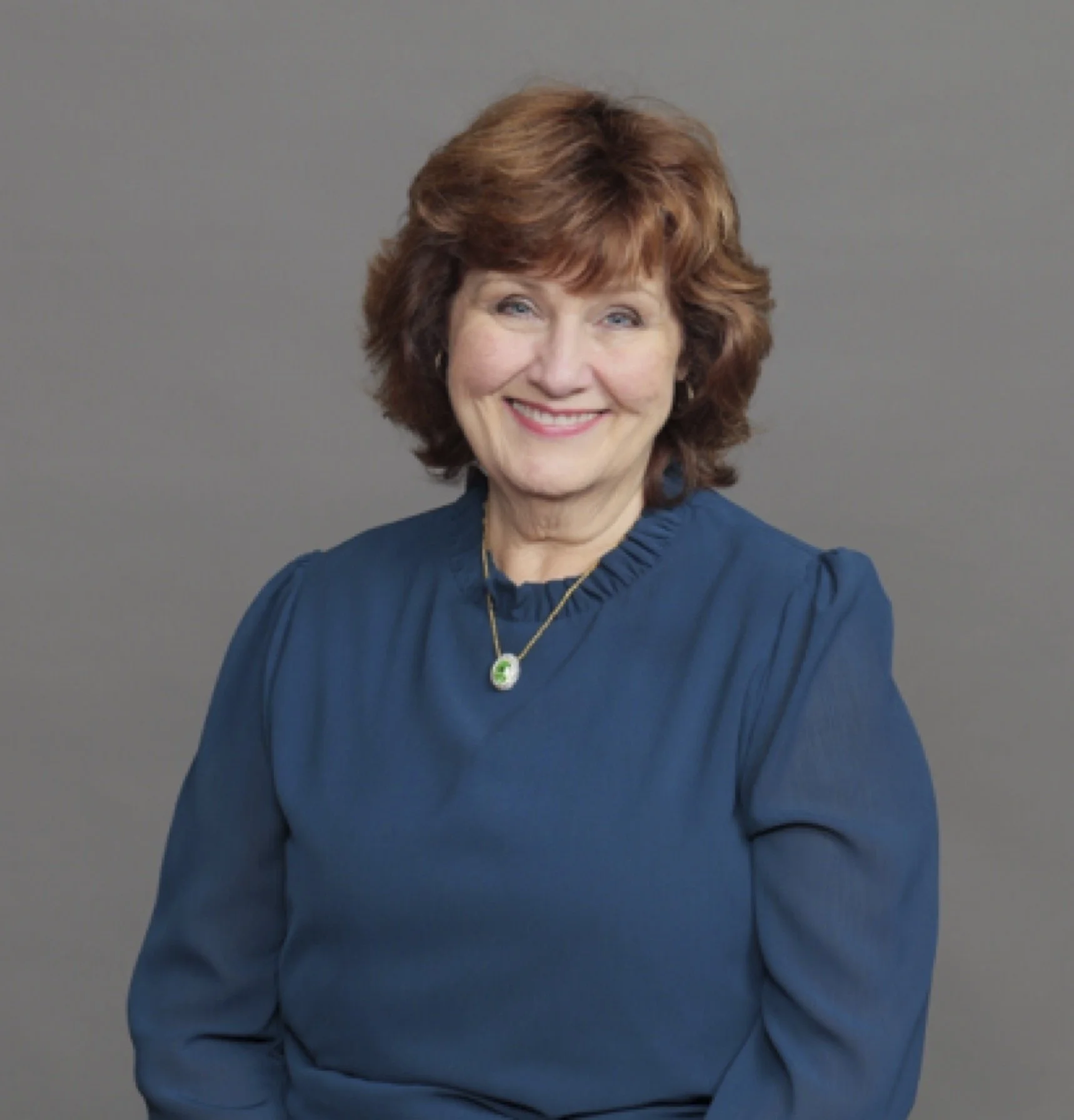 A woman with short, wavy brown hair, smiling, wearing a blue top and a silver necklace with a green pendant, against a plain gray background.