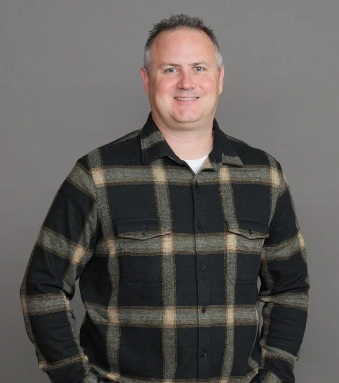 A man with short dark hair smiling, wearing a black and beige plaid button-up shirt, against a gray background.