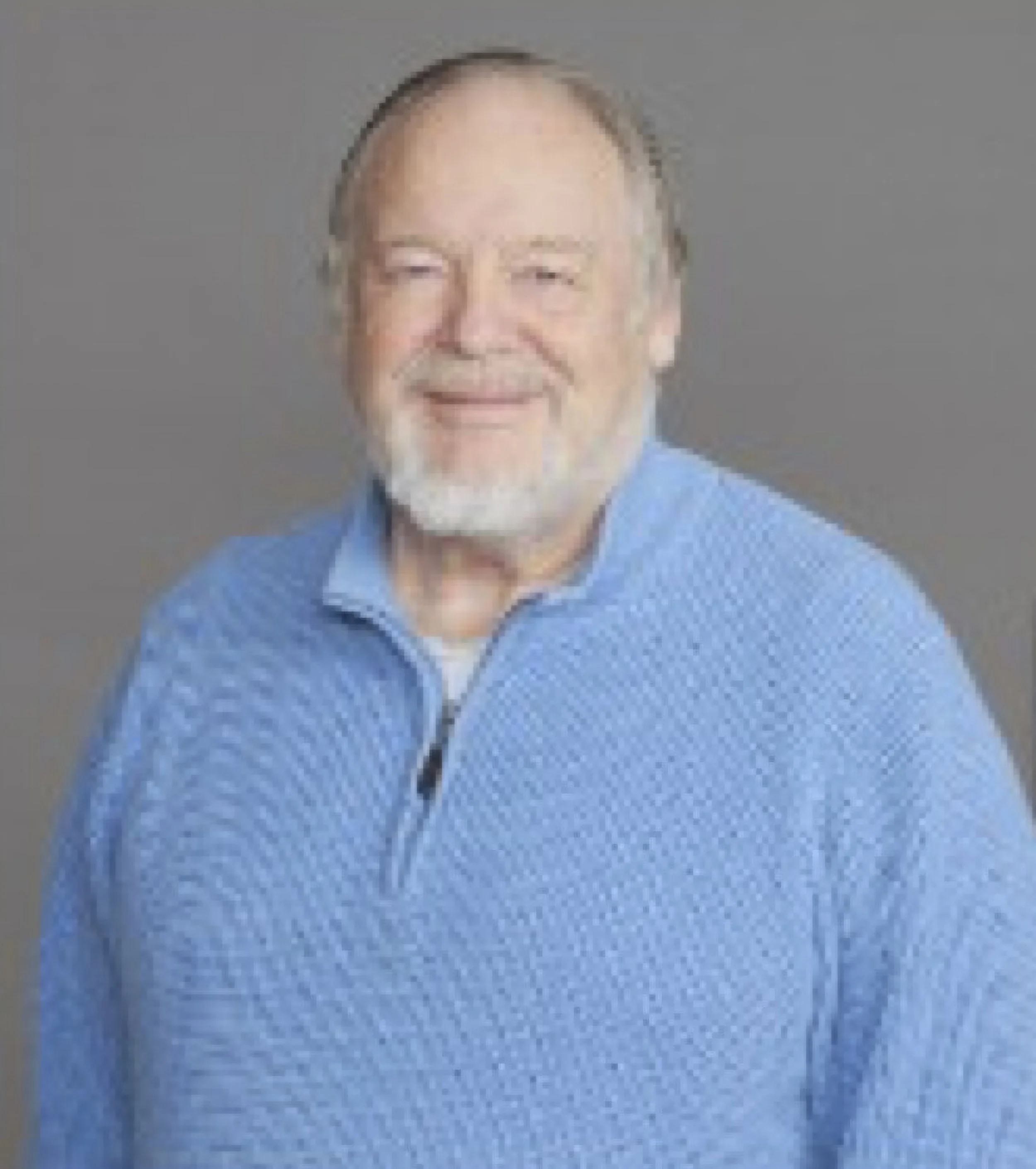 Portrait of a smiling older man with gray hair and beard, wearing a blue zip-up sweater, against a gray background.