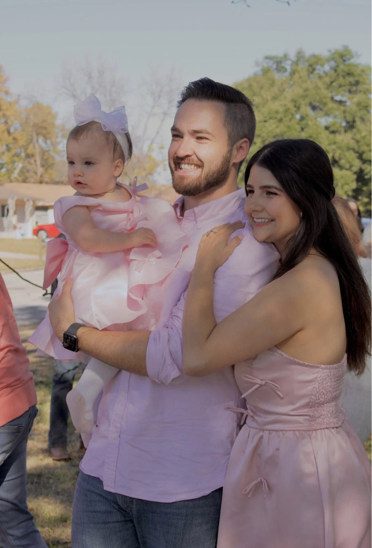 A smiling man holding a young girl dressed in pink, with a woman leaning on his shoulder, outdoors during daytime with trees in the background.