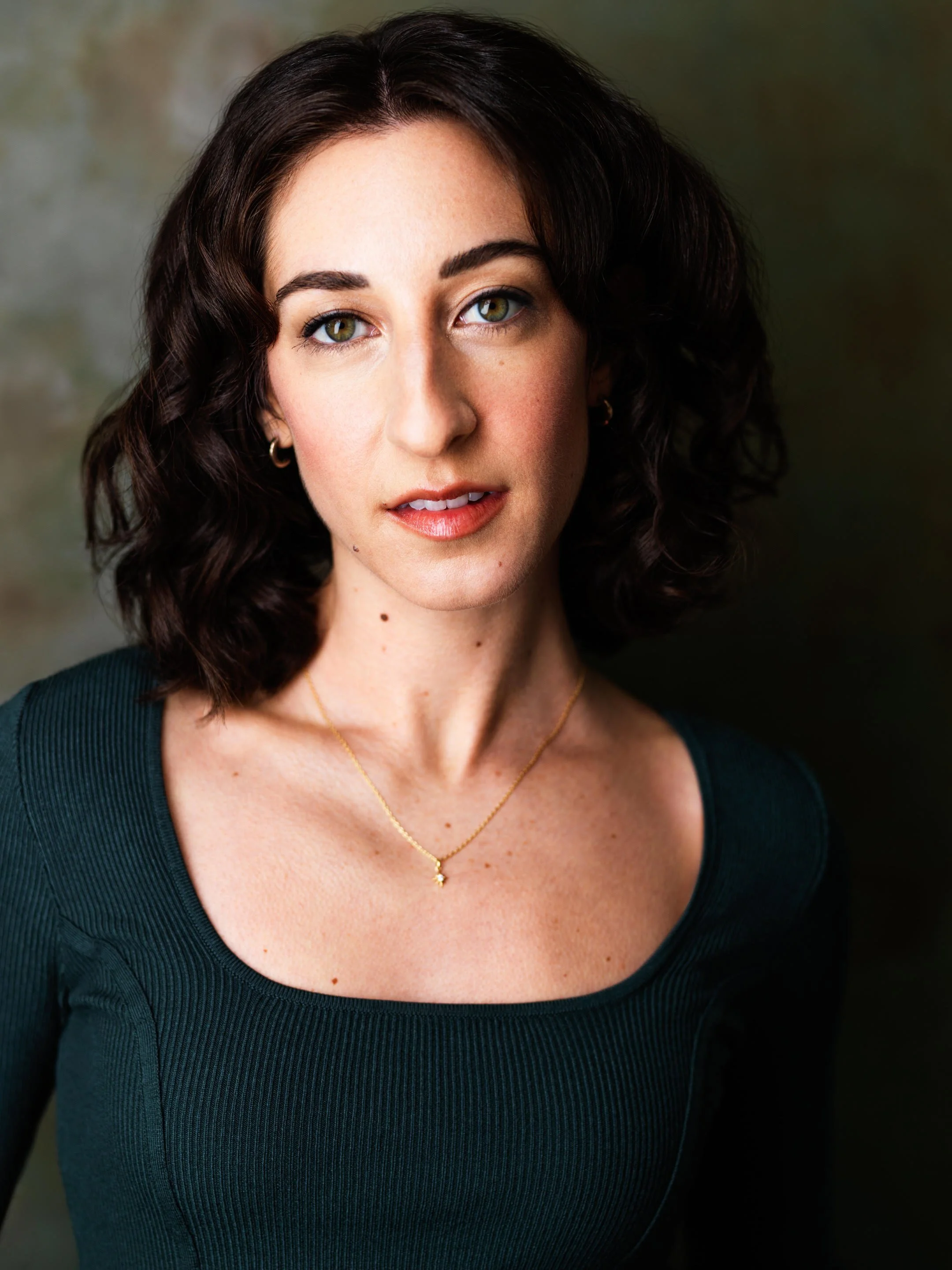 Close-up portrait of a woman with dark curly hair, green eyes, wearing a black top and gold jewelry, against a dark, blurred background.