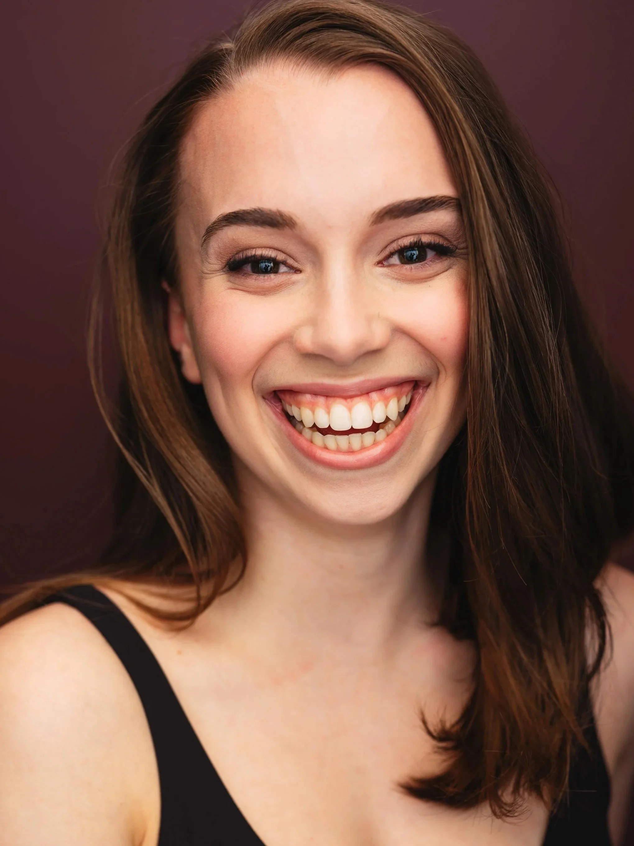 Close-up of a young woman with brown hair smiling, wearing a black sleeveless top, against a purple background.