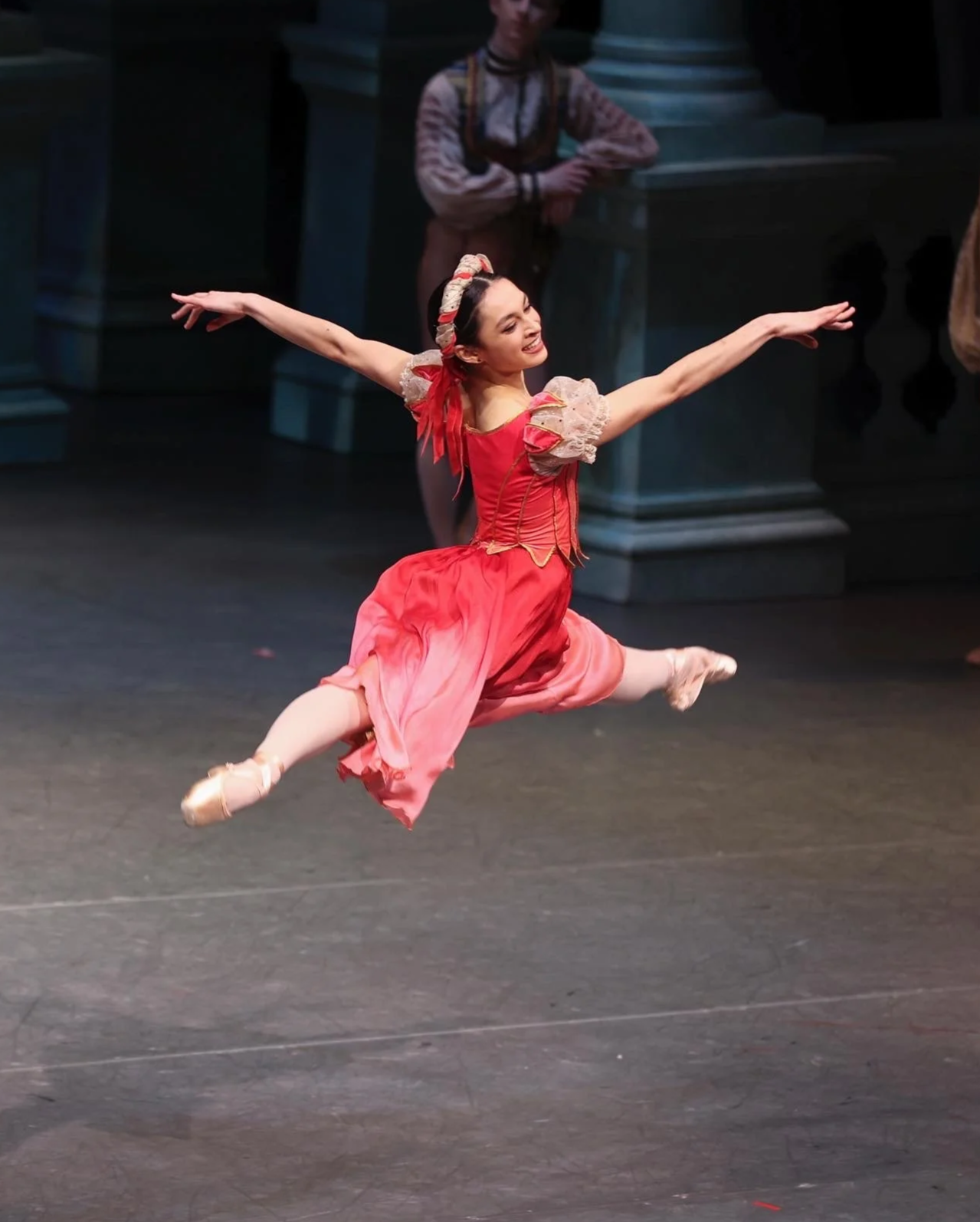 A young ballet dancer in a pink and red costume performing a dance leap on stage, with a person in period costume in the background.