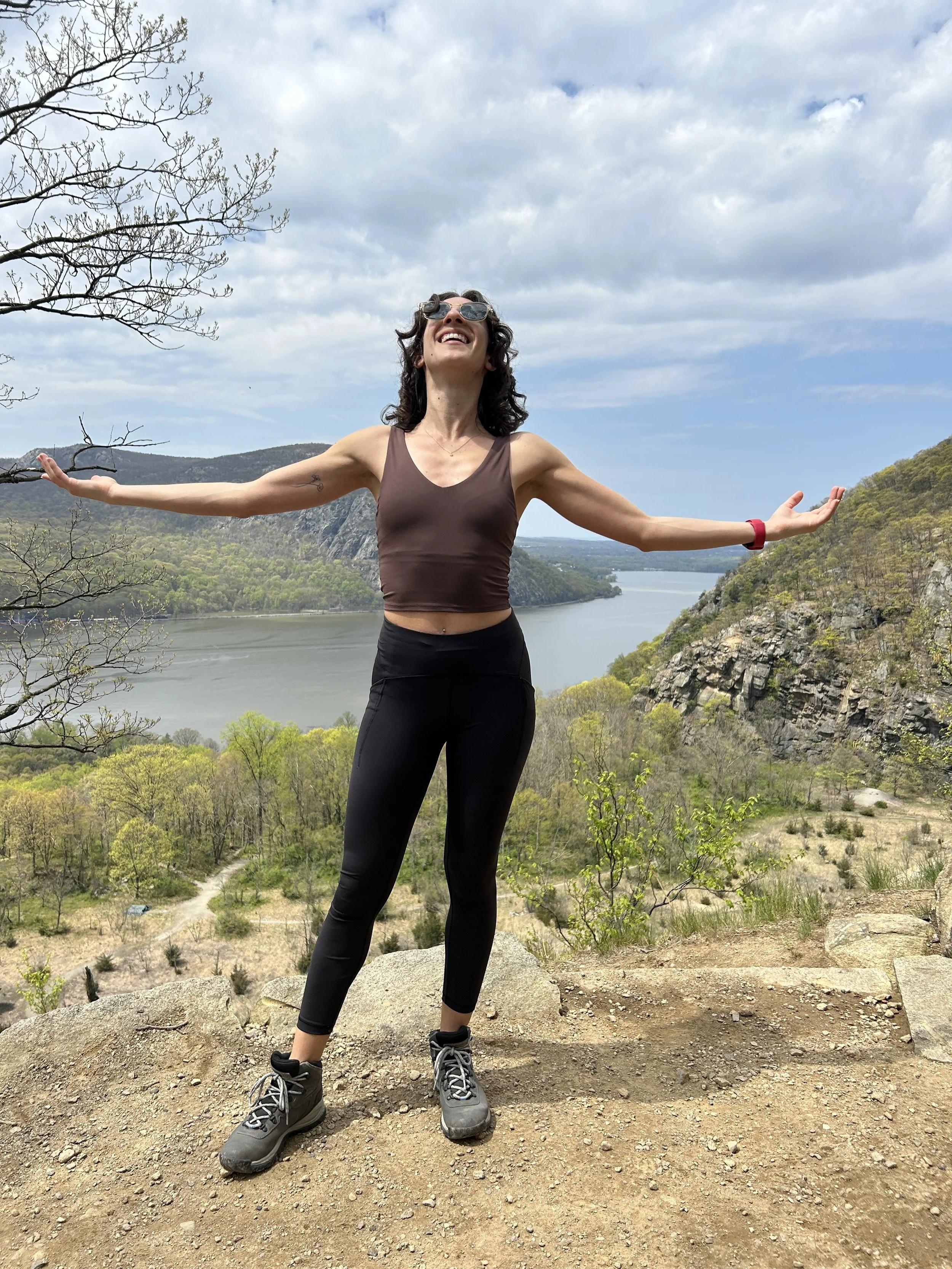 A woman standing on a dirt trail with arms outstretched, smiling and looking up, with a river and mountains in the background under a partly cloudy sky.