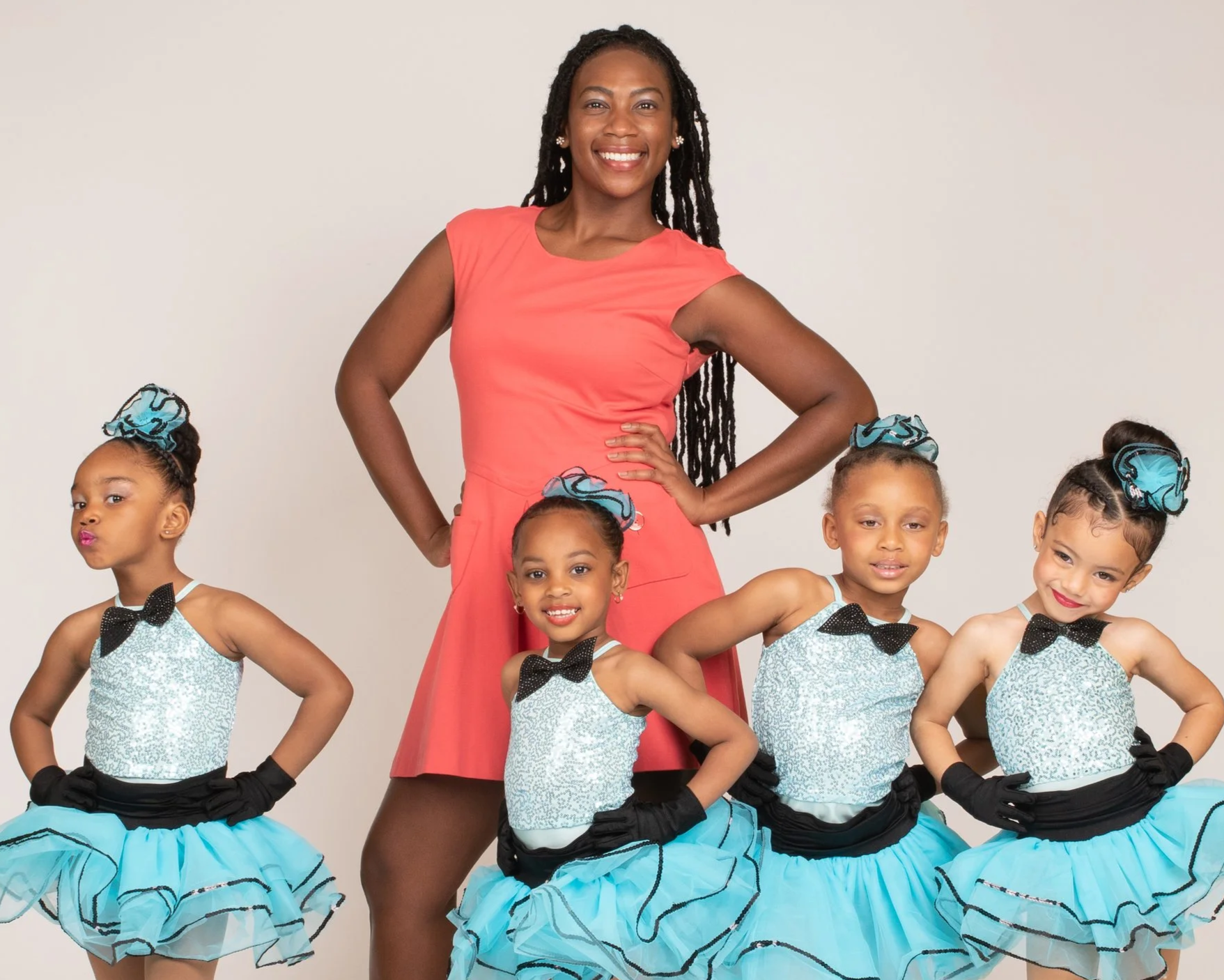 A smiling woman in a coral dress standing with her hands on her hips, surrounded by four young girls in matching dance costumes with blue skirts, white sequined tops, black gloves, and blue hair accessories, all posing confidently.