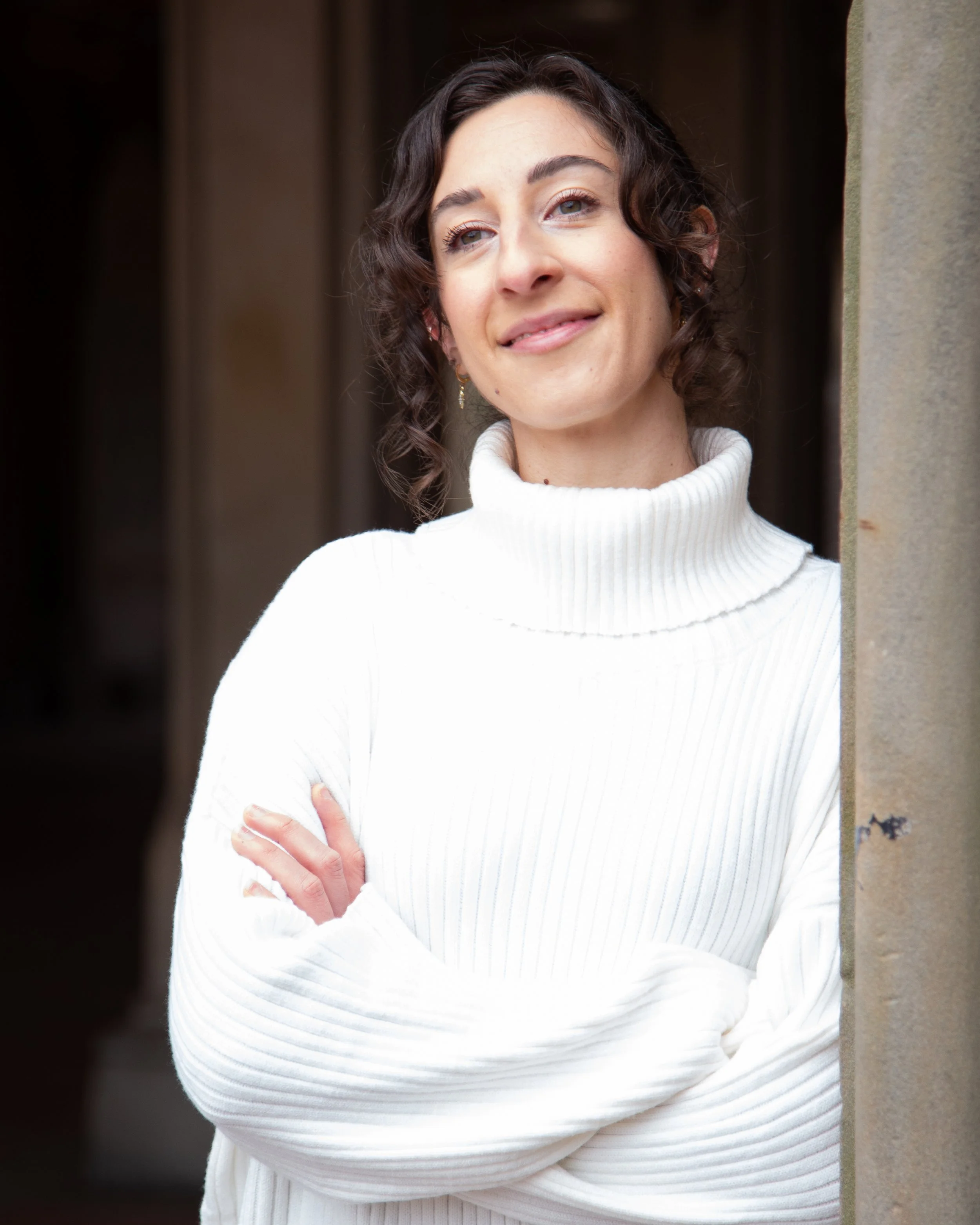 Woman with dark curly hair wearing a white turtleneck sweater, smiling with arms crossed, leaning against a stone wall.