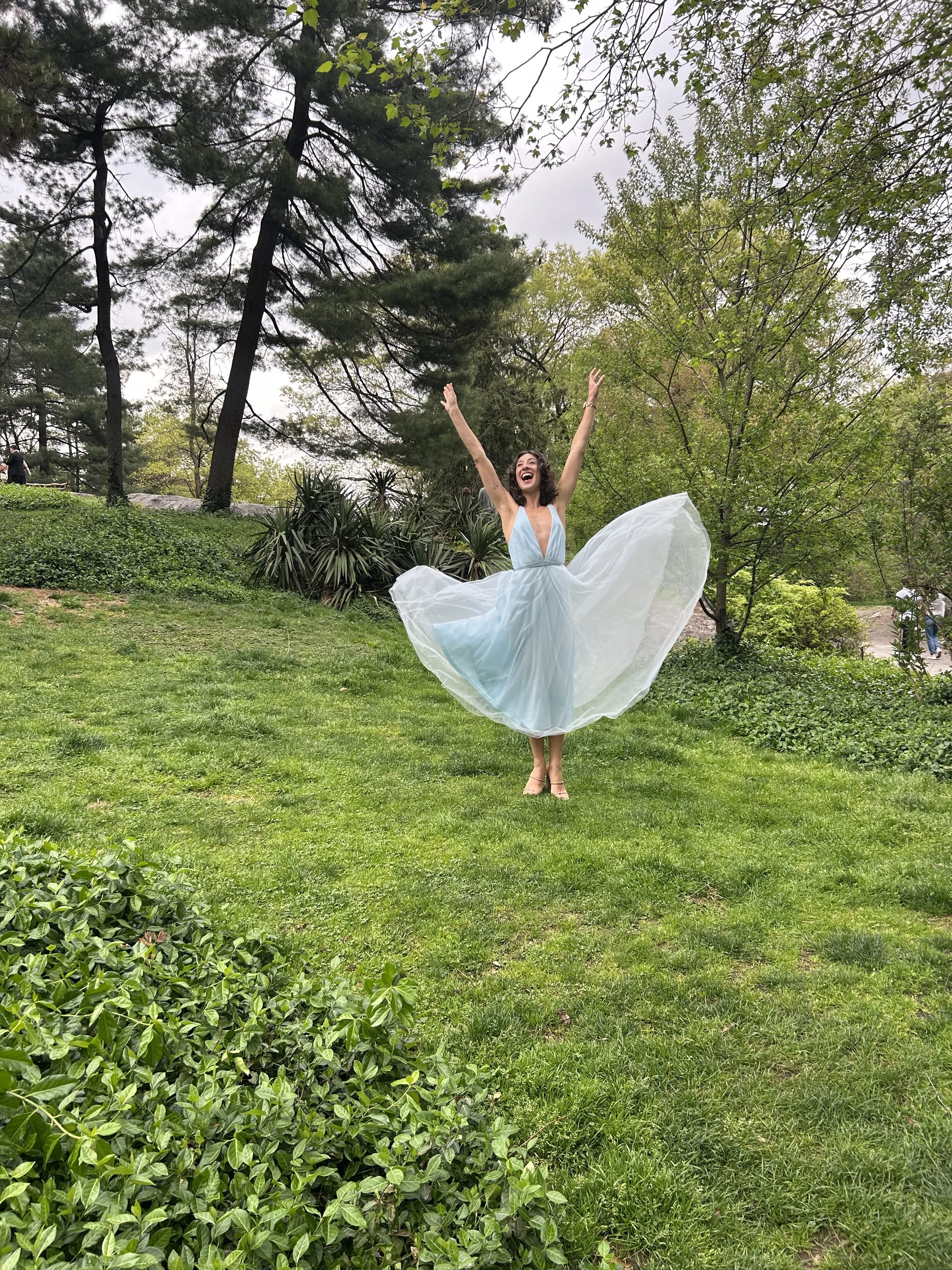 A woman in a light blue dress joyfully jumping in a green park with trees, bushes, and grass.