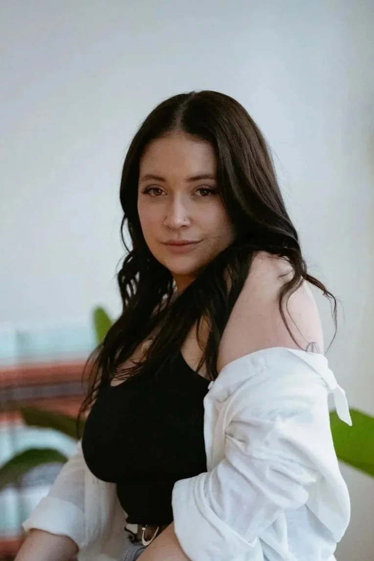 A woman with long dark hair and a nose piercing, wearing a black top and white jacket, standing indoors with trees and plants in the background.