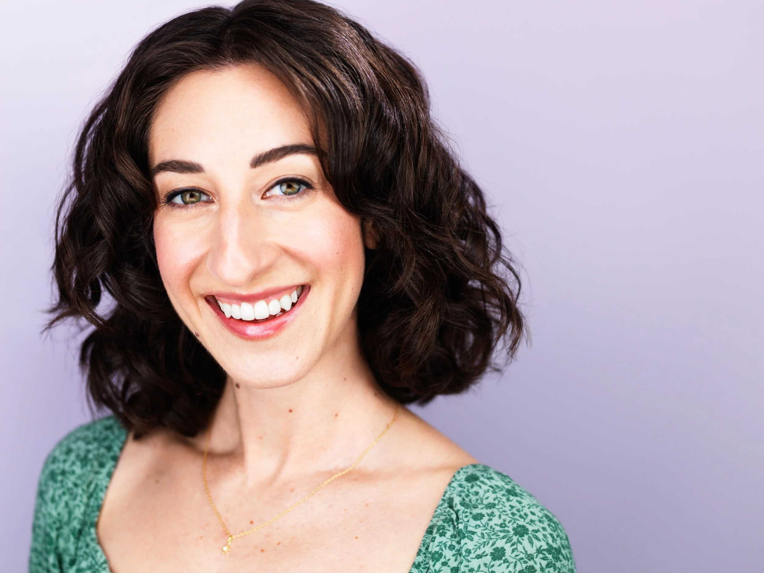 A woman with dark curly hair and fair skin smiling in front of a light-colored background, wearing a green patterned top and a gold necklace.