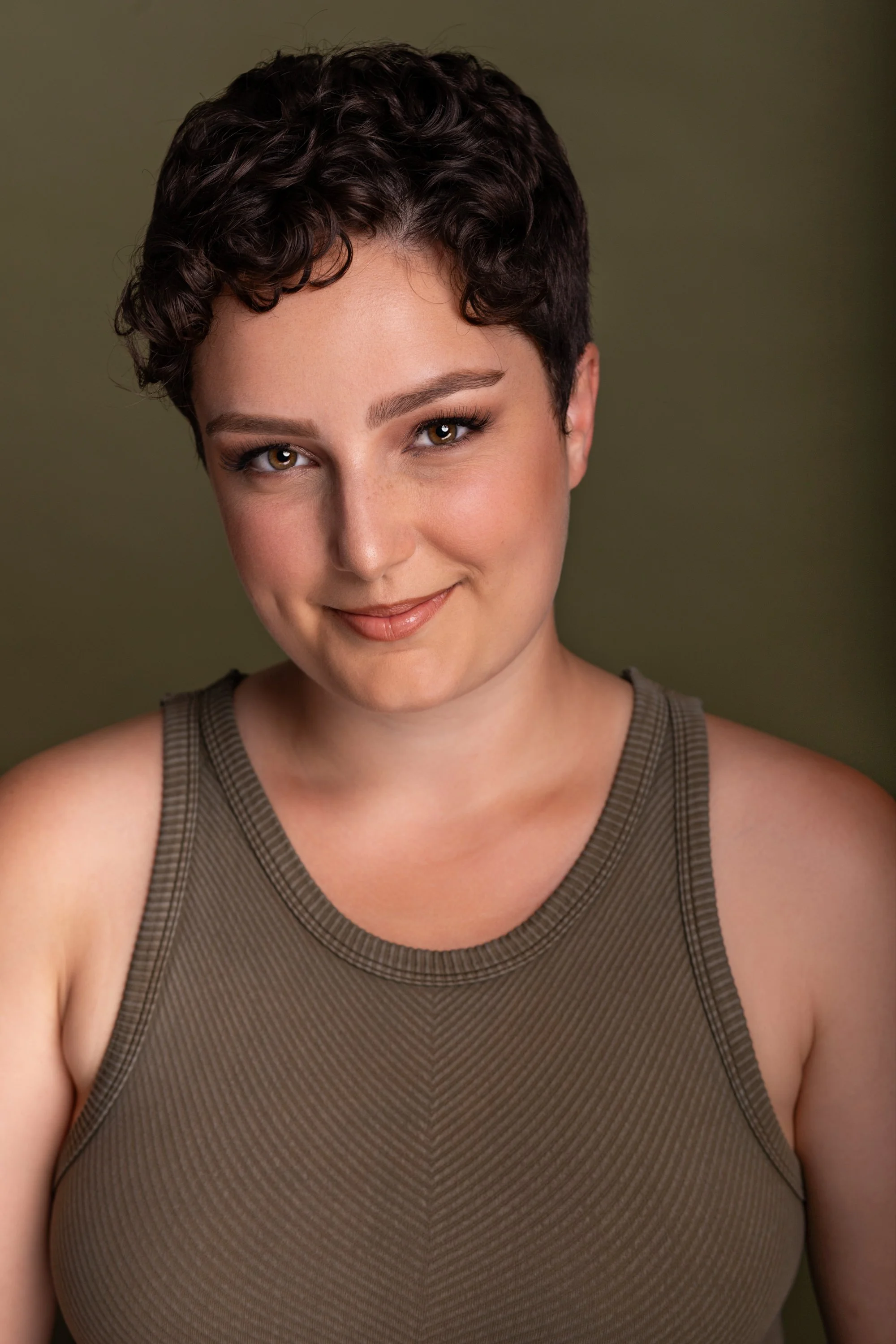 A young woman with short, curly dark hair and fair skin. She is wearing a sleeveless, olive-green top and has a subtle smile. The background is a muted green color.