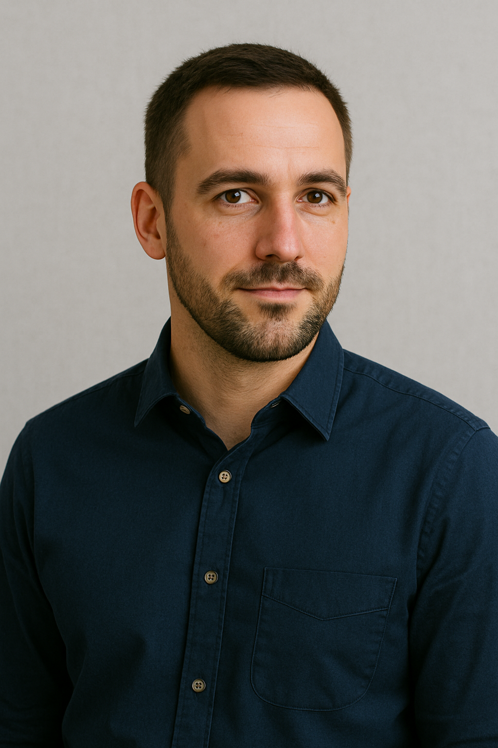 A young man with short brown hair and a beard, wearing a navy blue button-up shirt, looking at the camera with a slight smile, against a plain light gray background.