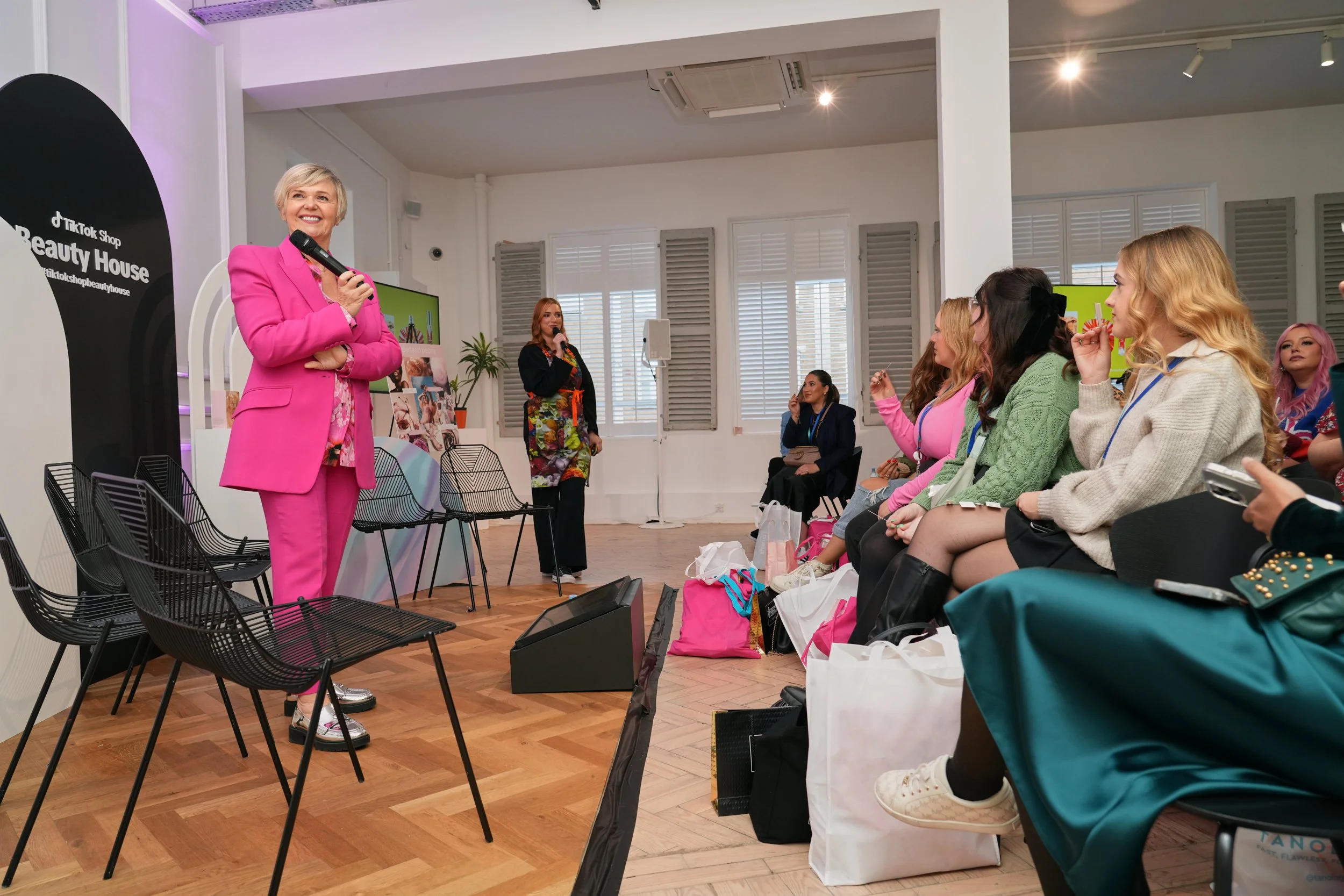 A woman in a bright pink suit holding a microphone and speaking to an audience in a well-lit room with white walls and large windows. The audience, mostly young women, is seated and listening attentively. There are four empty chairs in the front, and several bags and belongings on the floor near the seated women.