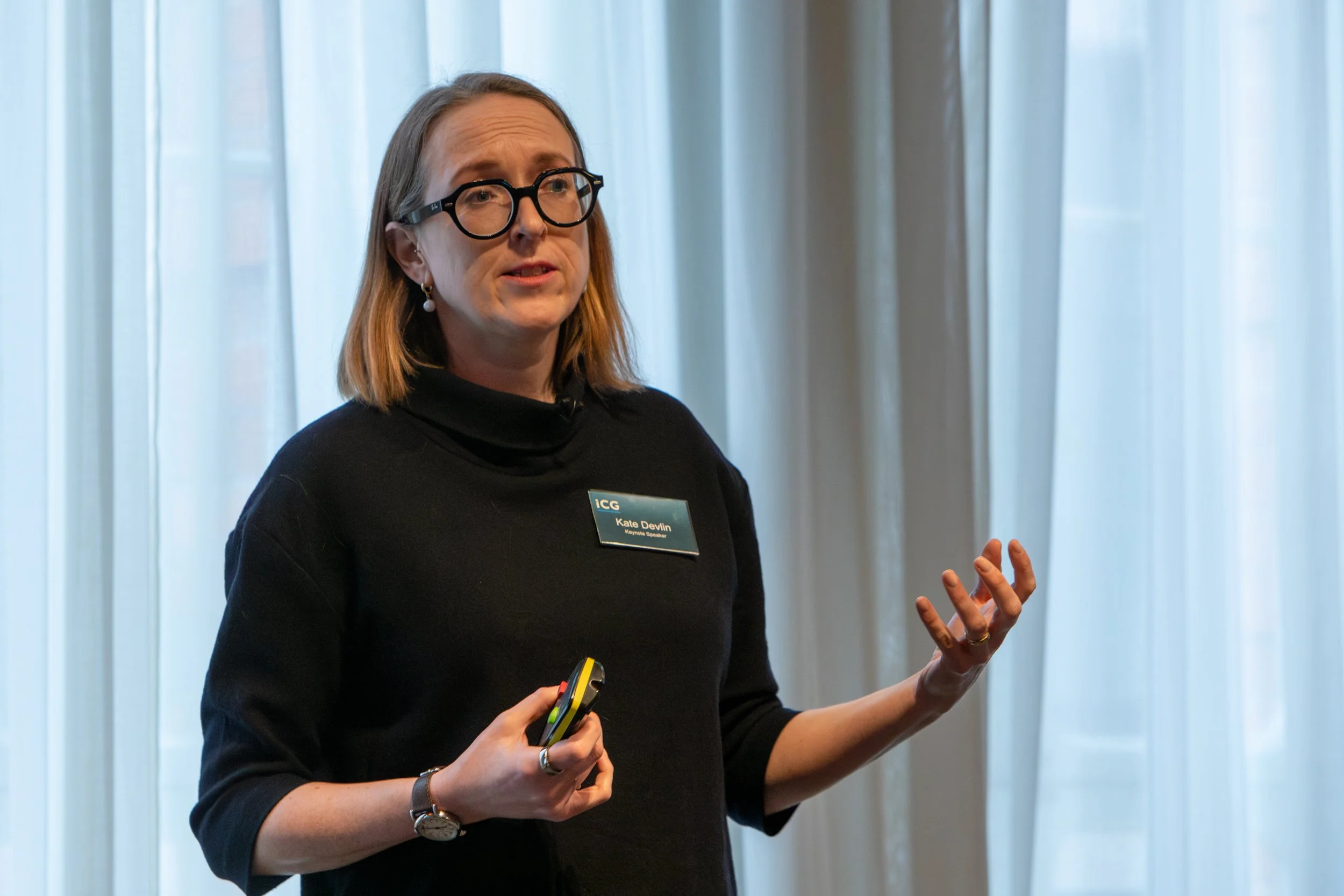A woman with glasses, wearing a black turtleneck and a name tag, speaking and gesturing during a presentation in a room with light curtains.