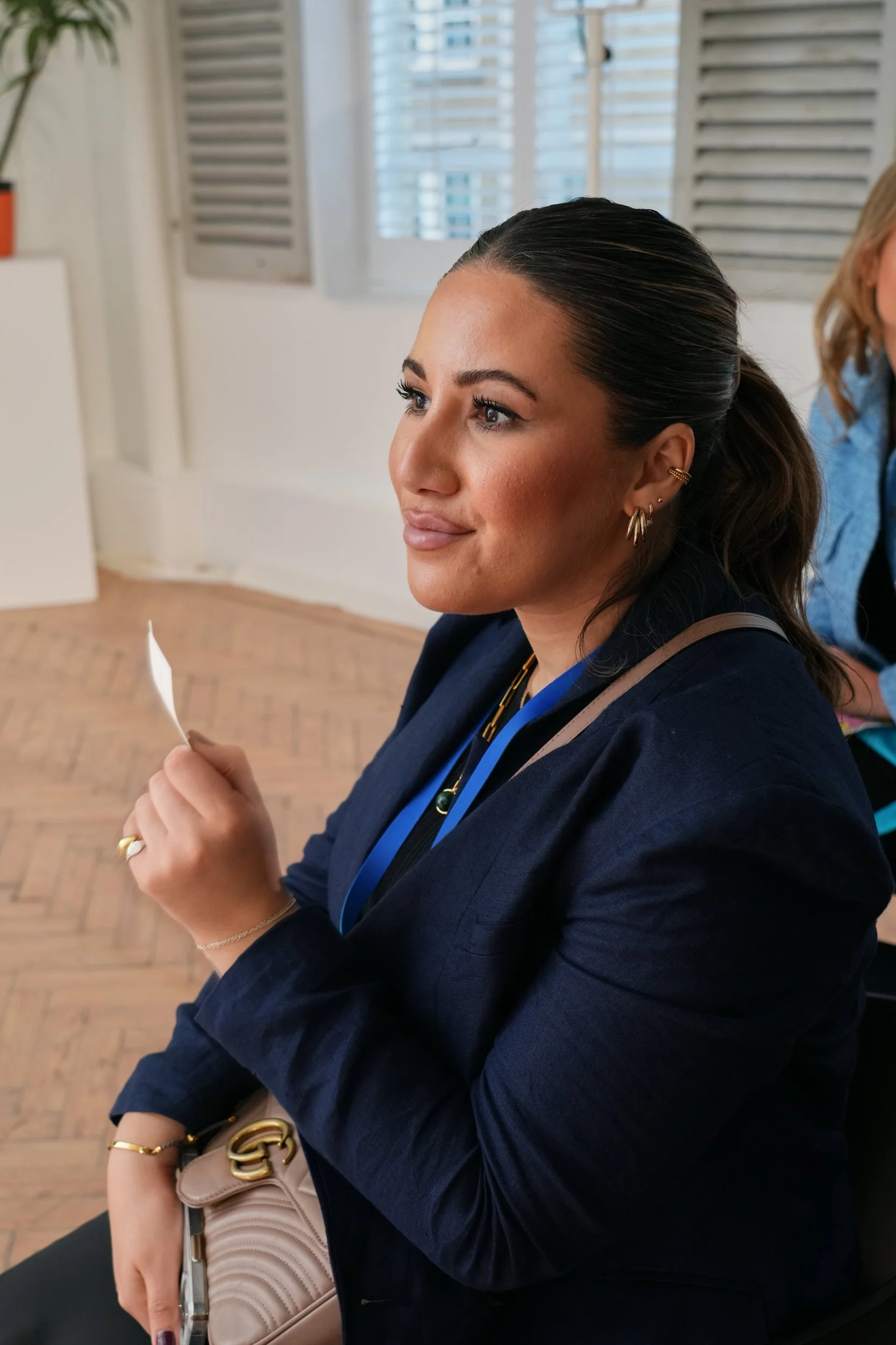 A woman with dark hair pulled back, wearing a navy blazer, gold earrings, holding a piece of paper, sitting indoors near a window with white shutters.
