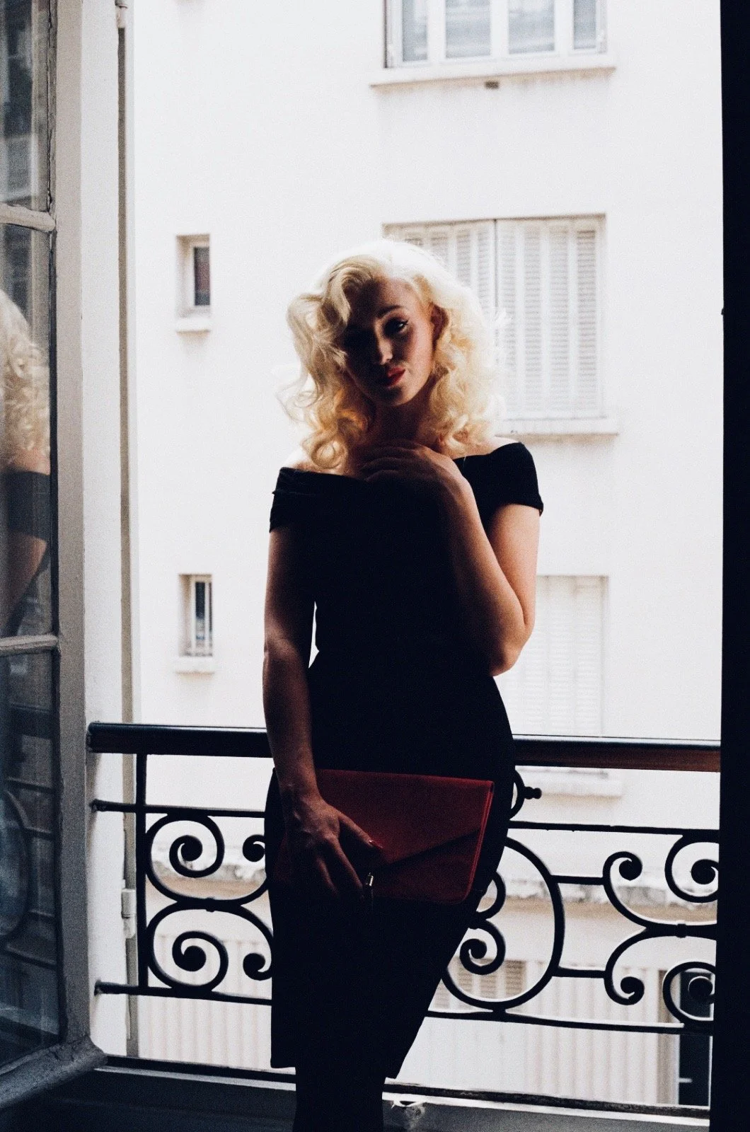 A woman with platinum blonde, voluminous curls standing on a balcony with ornate iron railing, wearing a black off-the-shoulder dress, holding a red clutch, during daylight with an urban building in the background.