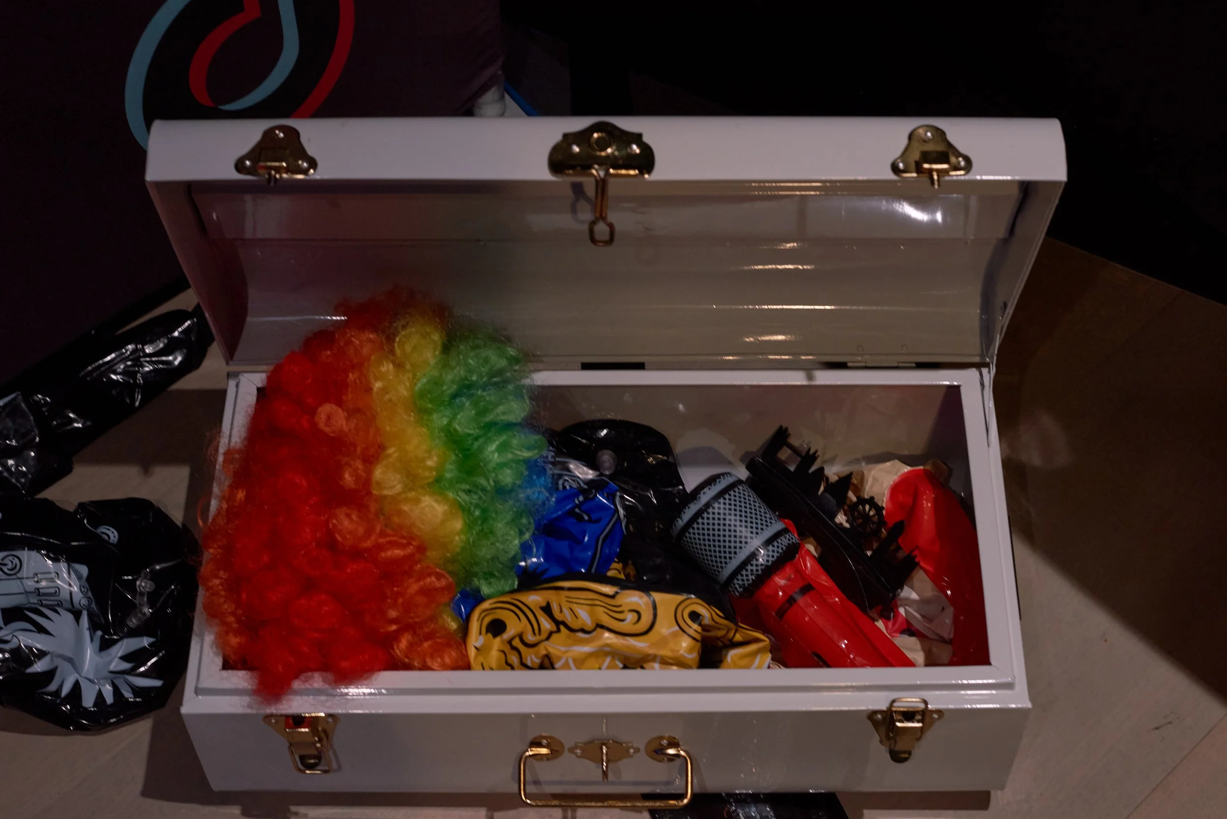 Open white box containing rainbow-colored feather boas, a red item, and other party supplies, with black plastic bags nearby.