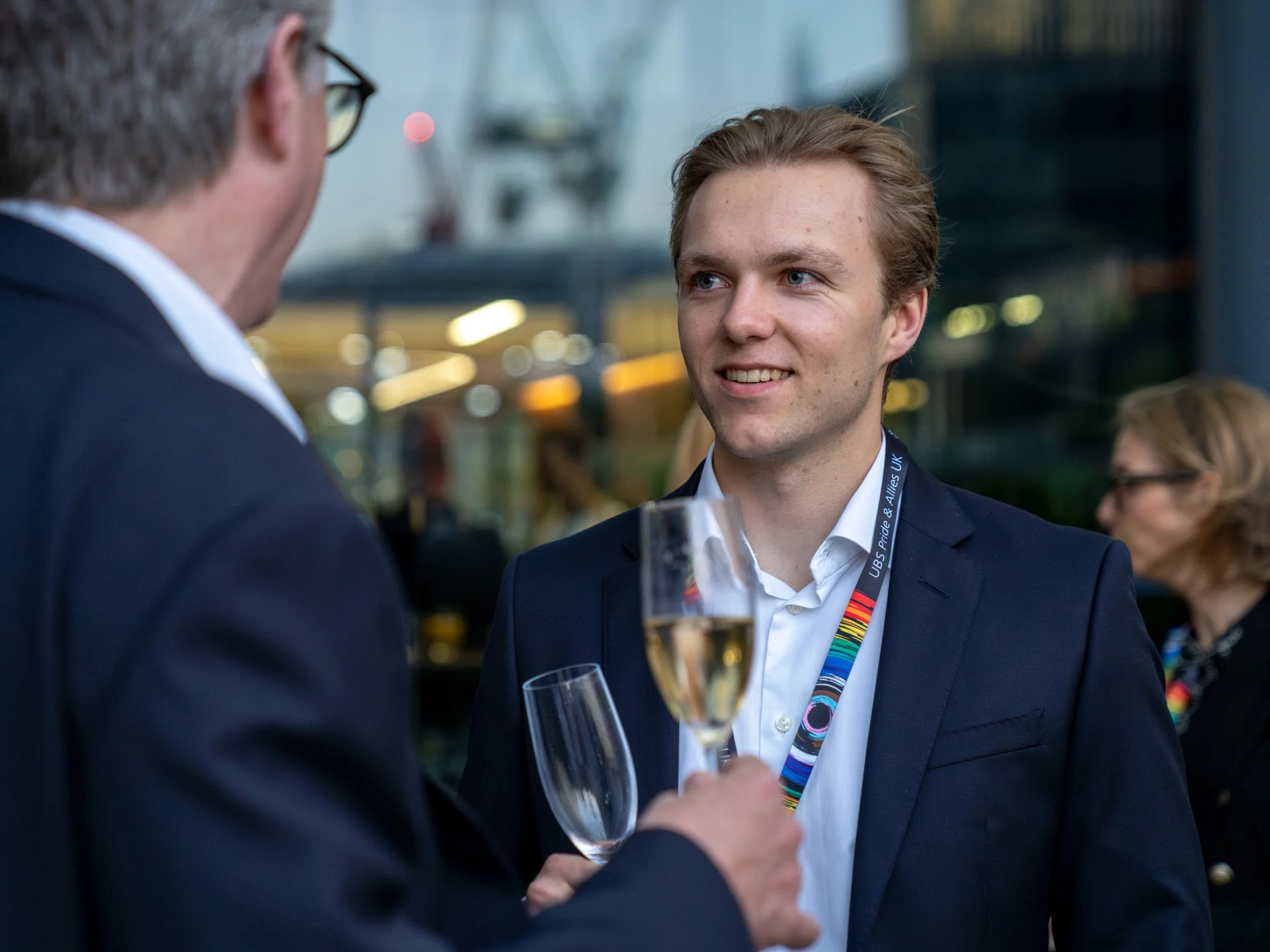 Two men in suits engaged in conversation at a social event, holding champagne glasses, with a blurred background of a building and other people.