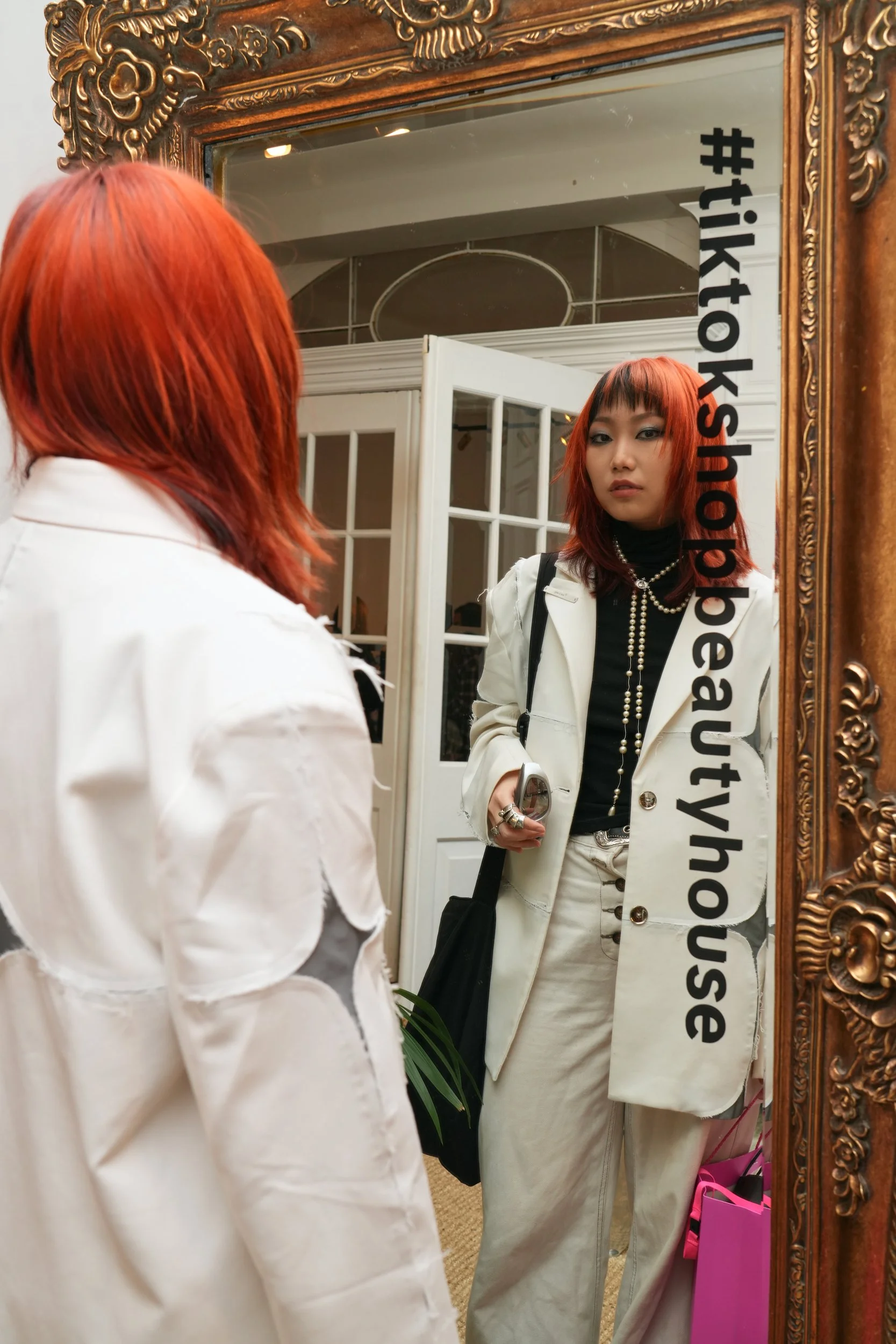 A young woman with red hair looking at her reflection in a large ornate gold-framed mirror. She is wearing a white blazer, black turtleneck, layered pearl necklaces, and cream-colored pants. The mirror has the hashtag '#tiktokshopoutyourself' written vertically on the right side.
