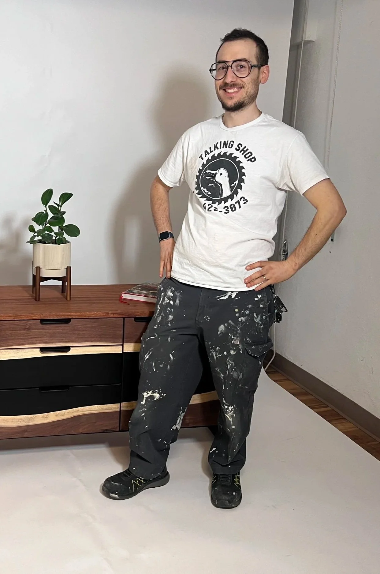 A man with glasses, a beard, and short hair is standing indoors on a white surface, smiling. He is wearing a white T-shirt that says 'Talking Shop' with a logo and phone number, black pants with paint splatters, and black shoes. His right hand is on his hip and his left hand rests on his thigh.