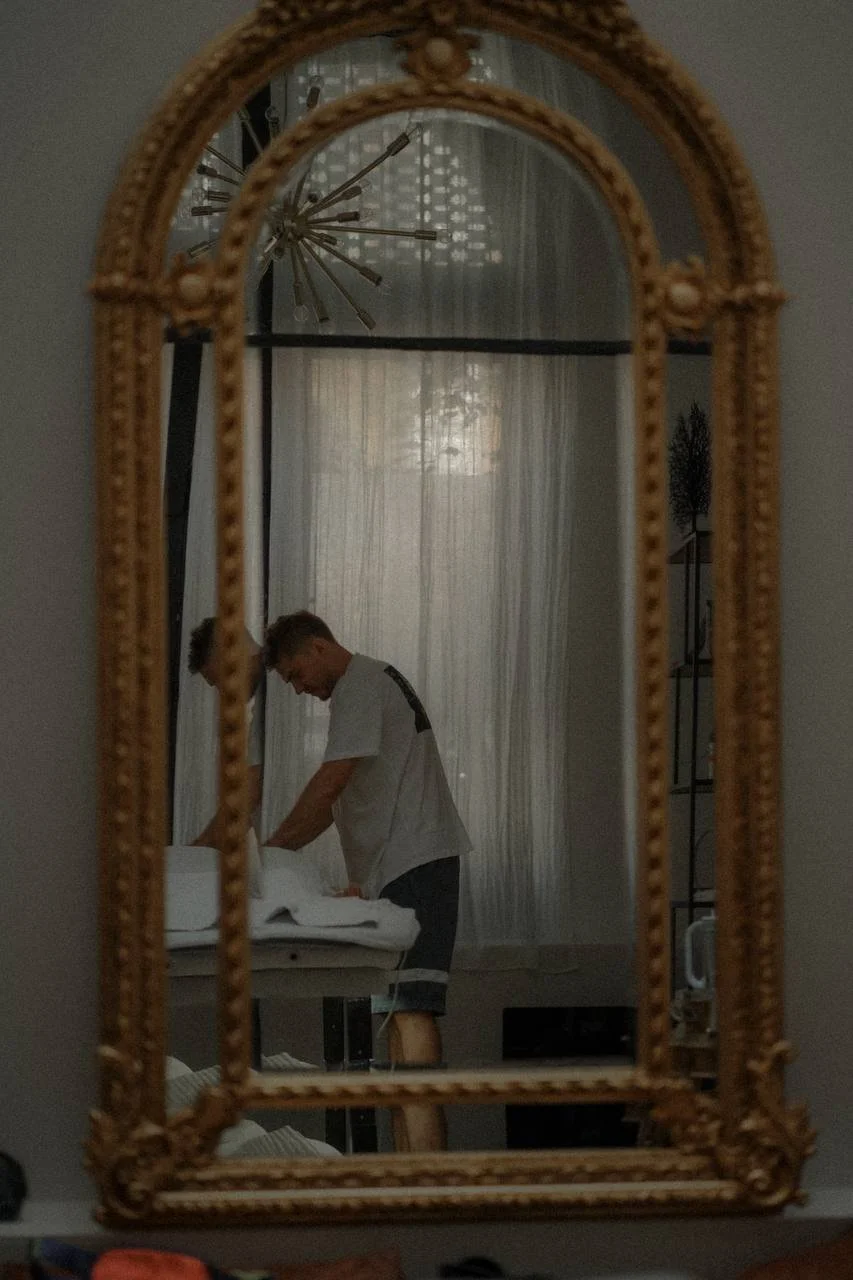 Reflection of a man making a bed, seen in an ornate mirror.