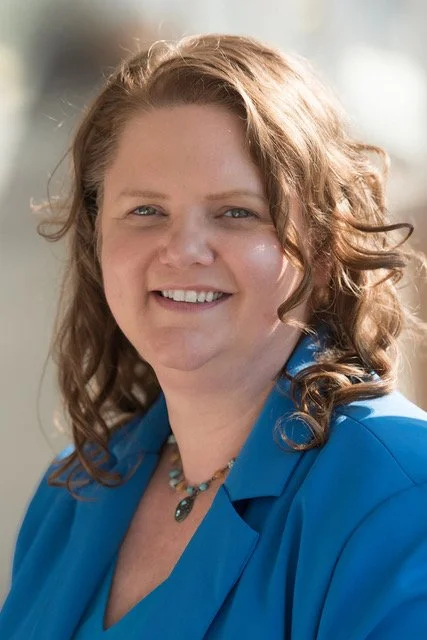 A woman with curly reddish-brown hair, smiling, wearing a blue shirt and a necklace, outdoors in sunlight.