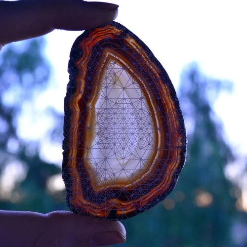 Hand holding a polished, cross-sectioned agate geode with glowing or highlighted interior layers and intricate pattern, with a blurred outdoor background.